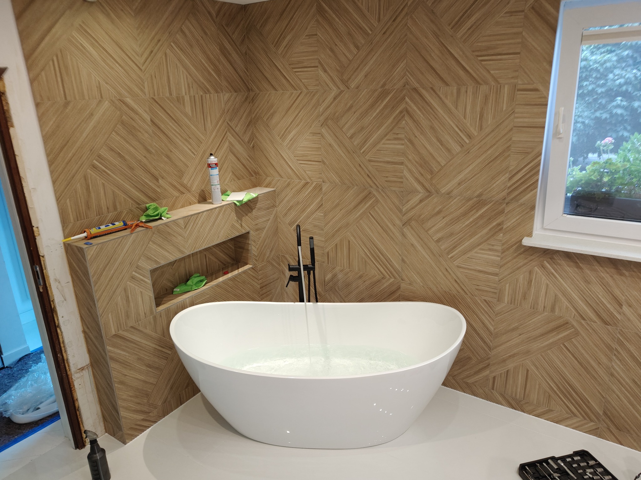 Nowoczesna łazienka z wolnostojącą wanną i ścianami wyłożonymi płytkami imitującymi drewno w geometryczny wzór. Widoczne narzędzia glazurnicze i chemia budowlana.