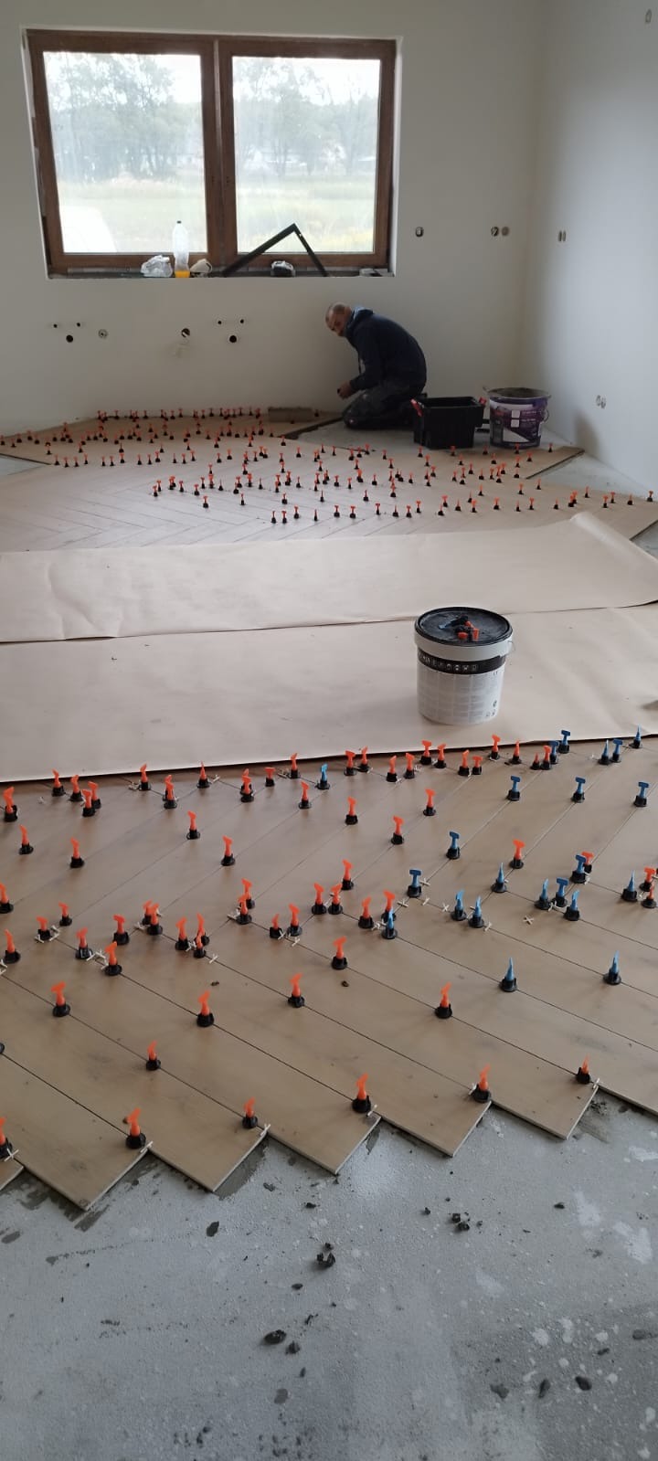 Układanie parkietu w jodełkę z użyciem systemu poziomowania płytek. Widoczny glazurnik i wiadro z klejem. Podłoga zabezpieczona papierem. Jasne wnętrze z oknem.