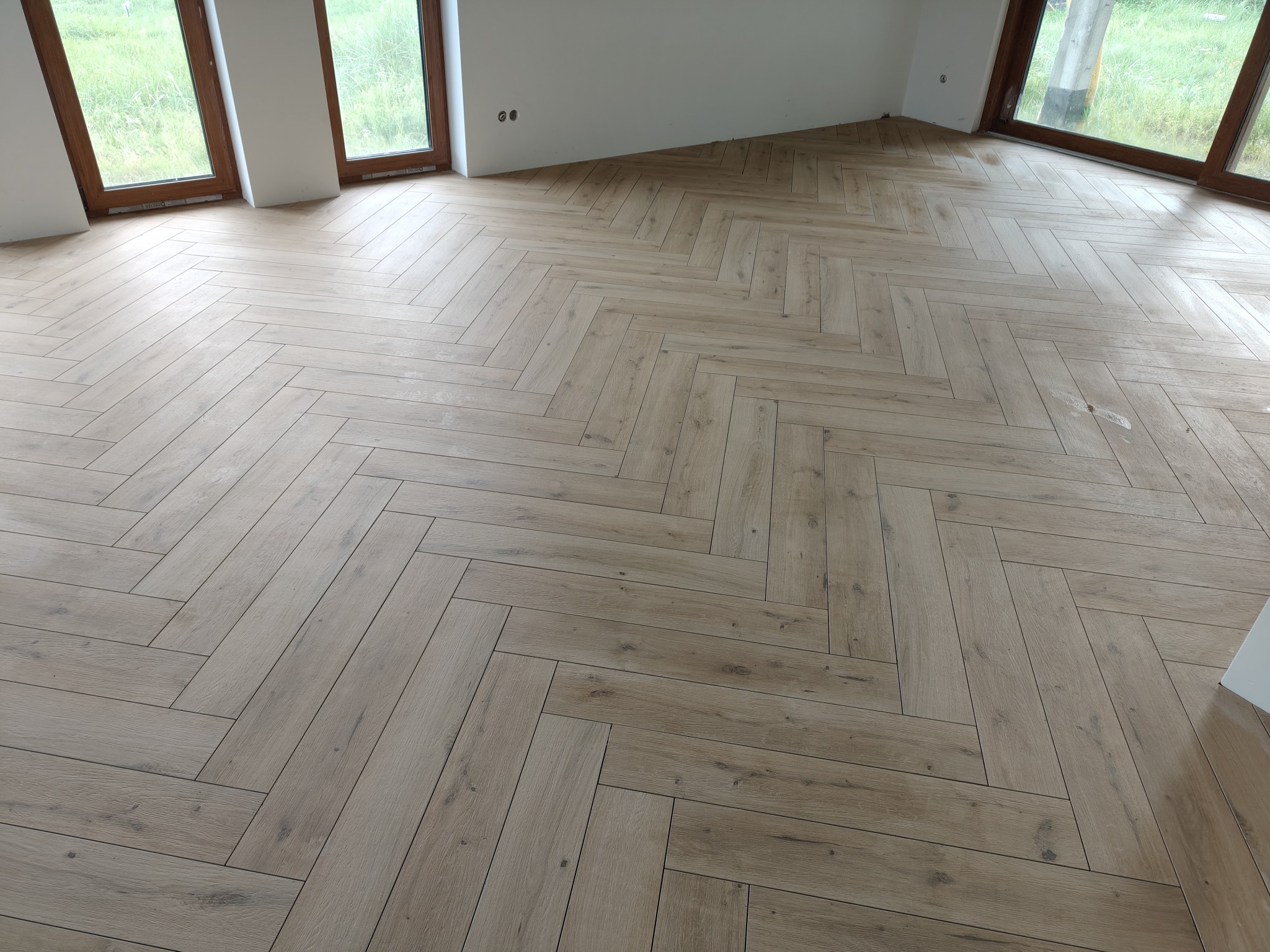 Jasne, drewnopodobne płytki ułożone w jodełkę na podłodze w nowym pomieszczeniu z oknami. Widoczne gniazdka elektryczne na ścianie.