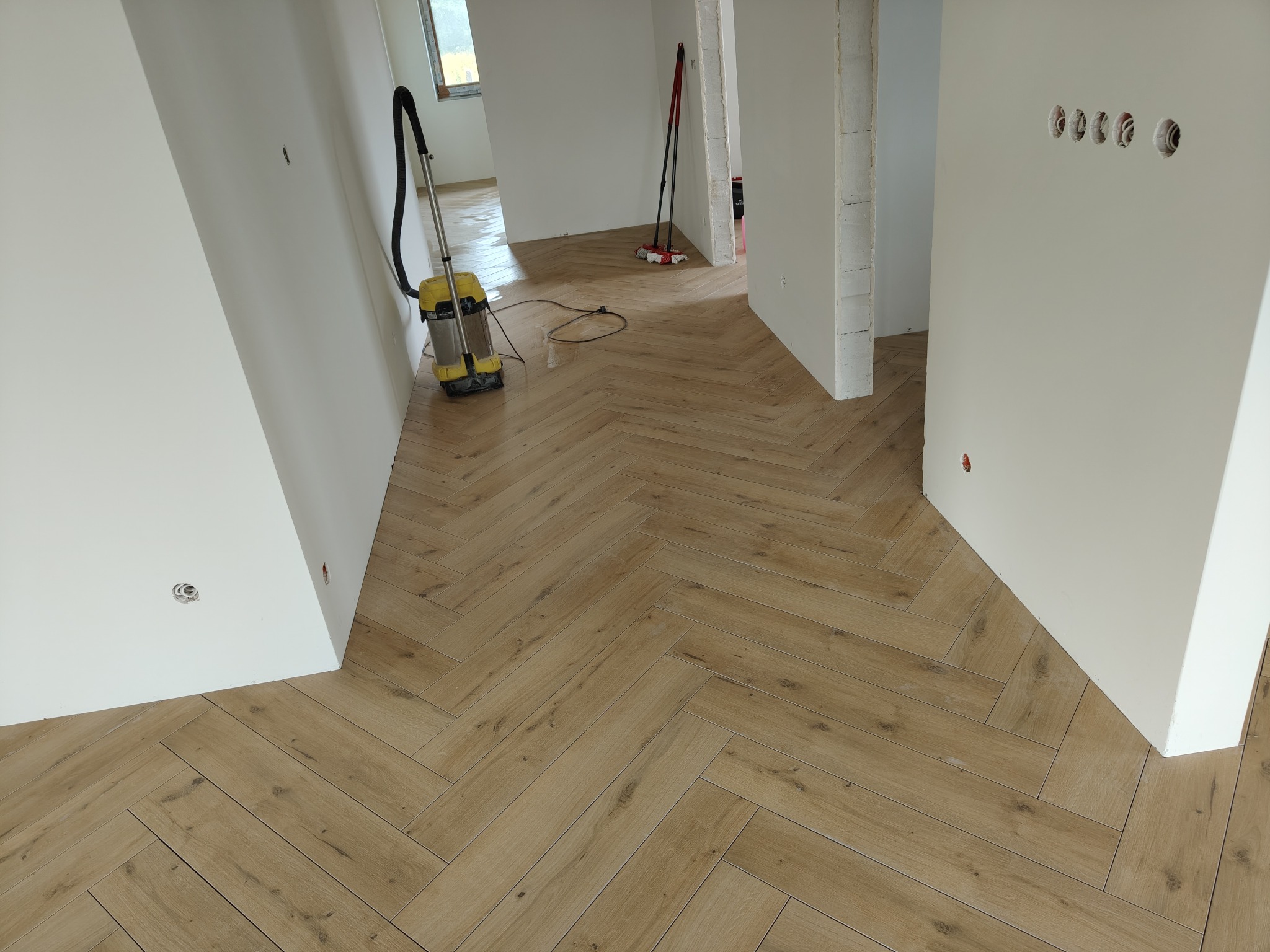 Ułożona podłoga z paneli w jodełkę w jasnym wnętrzu. Widoczny odkurzacz do wody i fragmenty ścian z instalacją elektryczną. Jasne, naturalne światło.