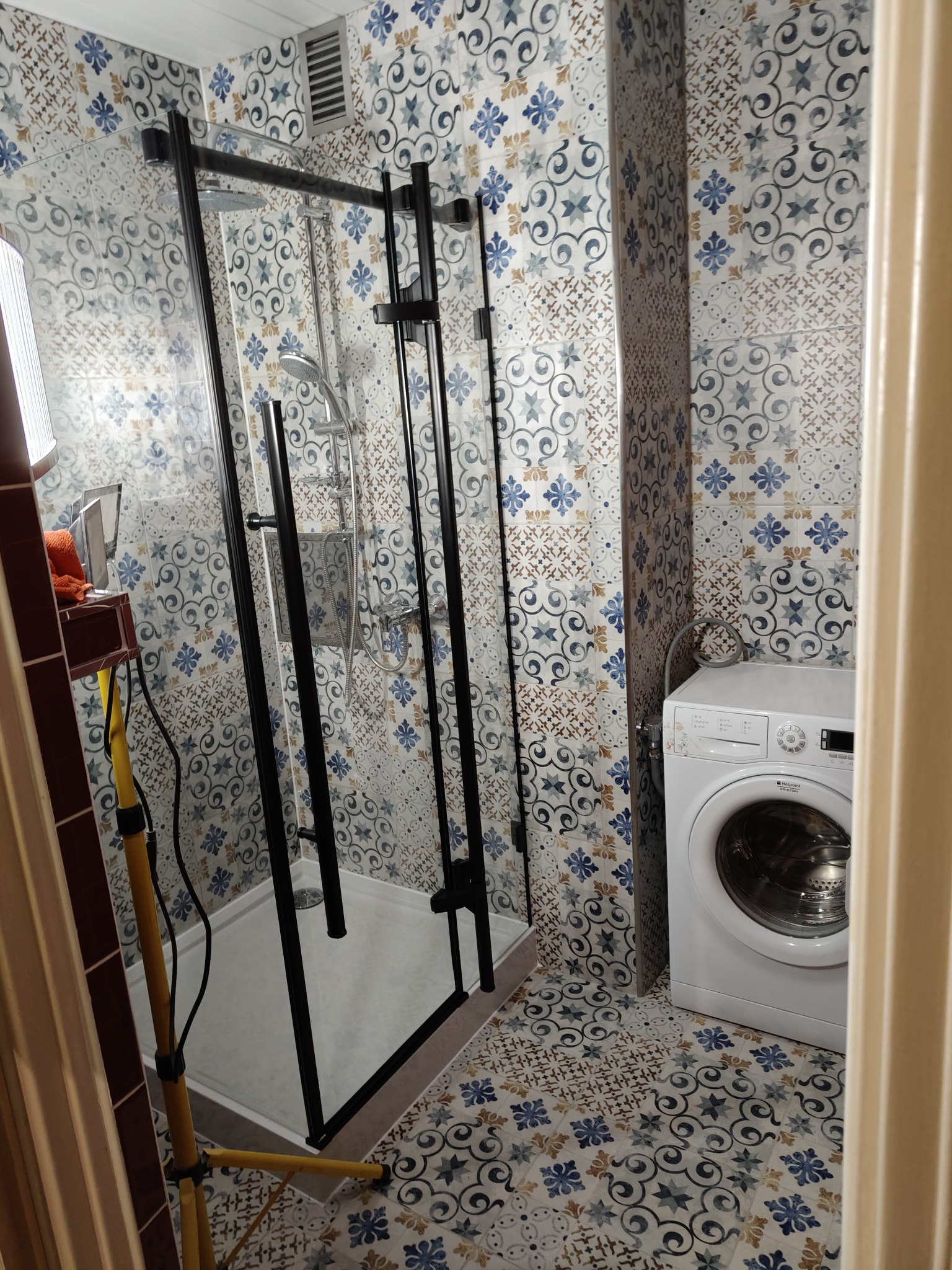 Wykończona łazienka z kabiną prysznicową typu walk-in z czarnymi profilami, wzorzystymi płytkami na ścianach i podłodze oraz pralką. Widoczny reflektor budowlany.