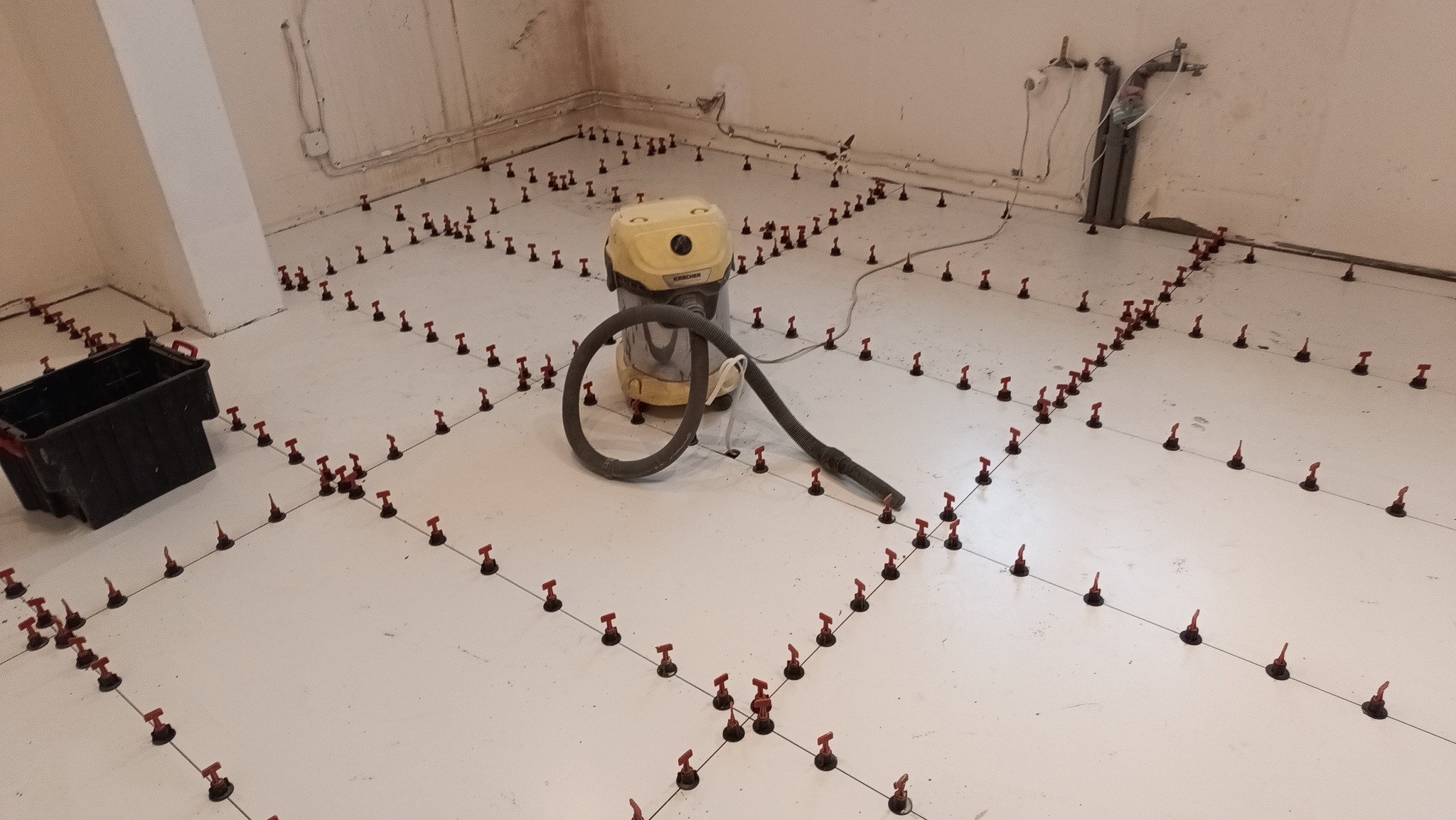 Biała podłoga z systemem poziomowania płytek, widoczny odkurzacz przemysłowy i czarny pojemnik. Przygotowanie do układania płytek, równomierne rozmieszczenie elementów.