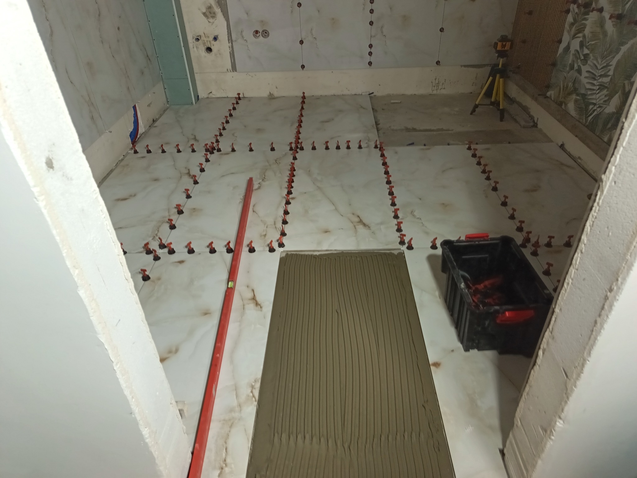 Układanie płytek podłogowych w łazience z użyciem poziomicy i systemu poziomowania. Widoczna nałożona warstwa kleju na płytce. W tle laser krzyżowy.