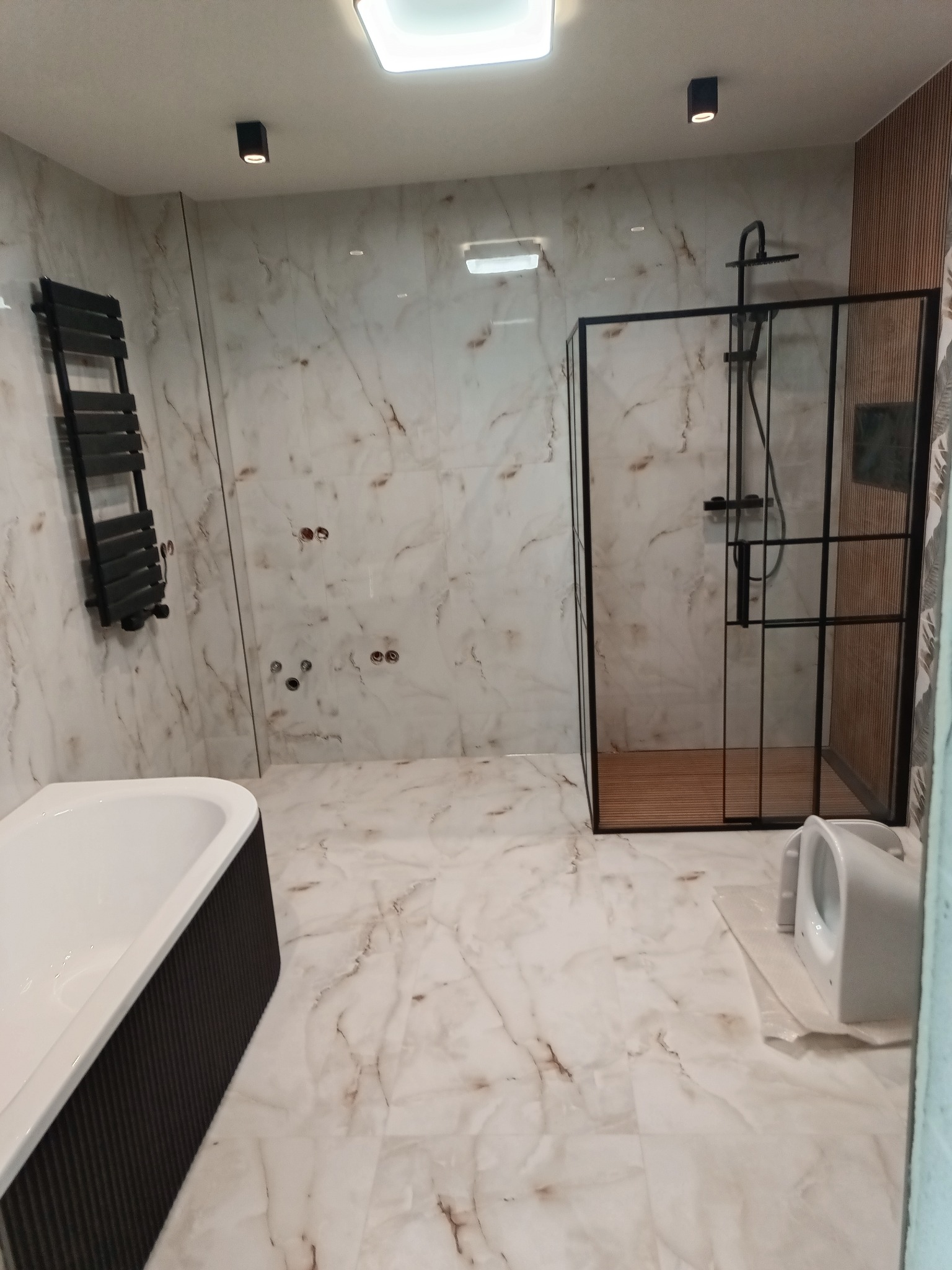 Nowoczesna łazienka z marmurowymi płytkami, czarną armaturą i kabiną prysznicową typu walk-in. Widoczna wanna z czarnym obramowaniem i grzejnik drabinkowy.