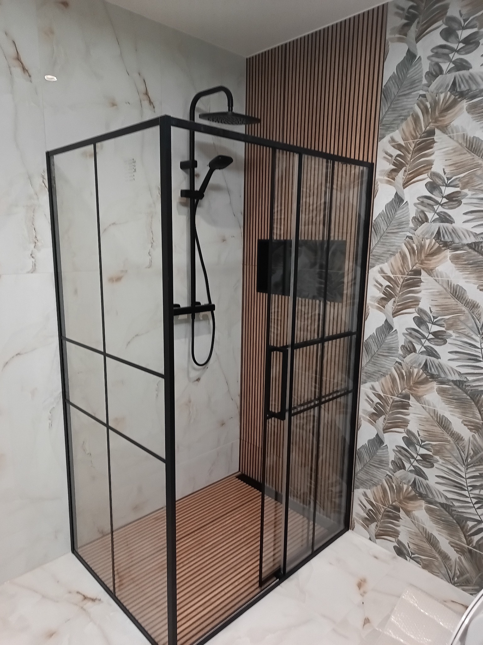 Nowoczesna kabina prysznicowa z czarnymi profilami na tle marmurowych płytek i drewnianych lameli. Widoczny deszczownica i wzorzysta tapeta z motywem liści.