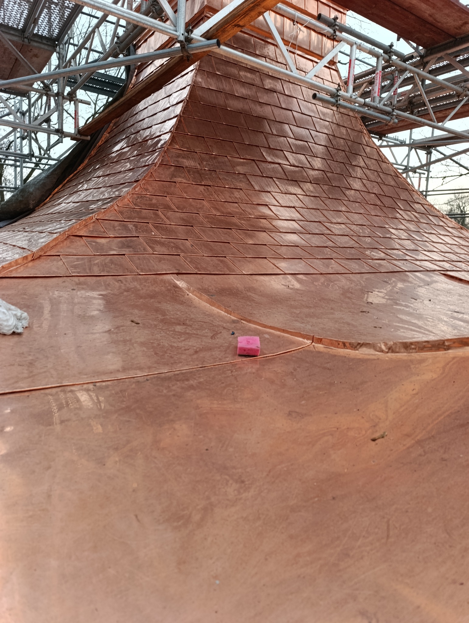 Miedziany dach w trakcie krycia, widoczne rusztowania i fragmenty blachy dachowej. Na gładkiej powierzchni leży różowa gąbka.