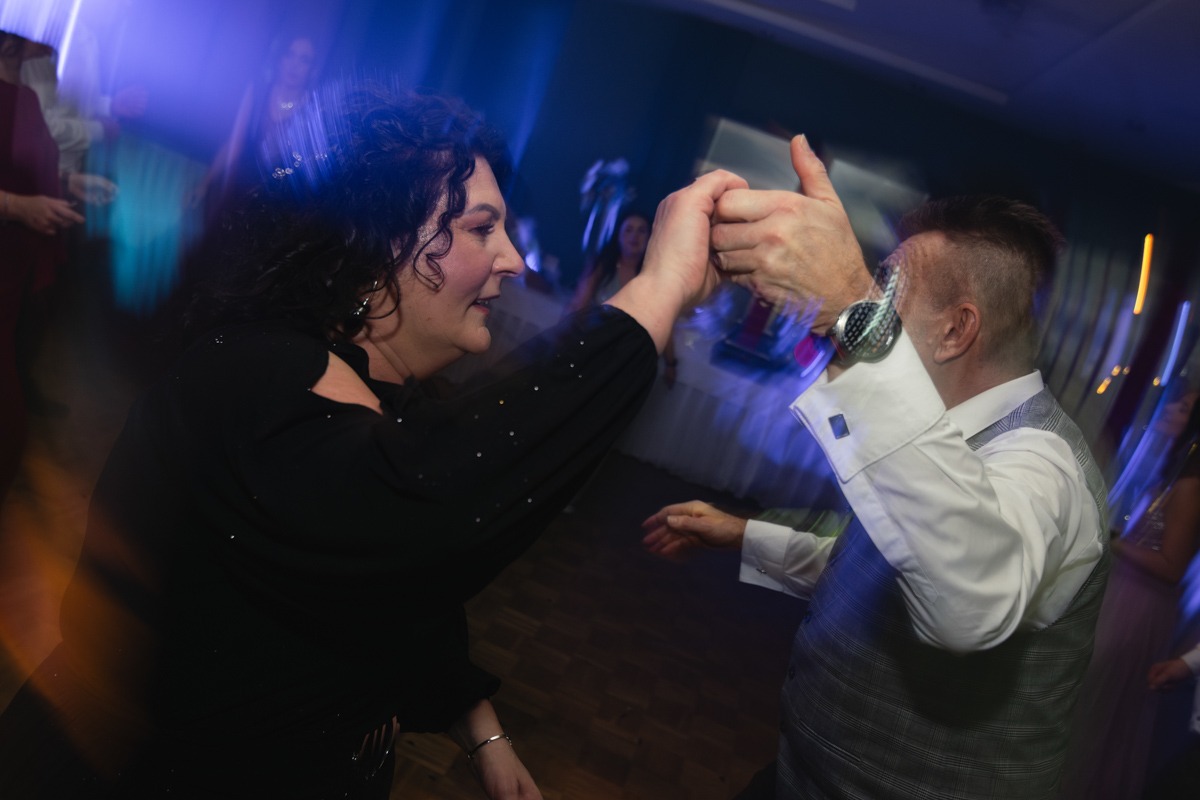 Para w tańcu podczas imprezy, kobieta w czarnej sukience z odkrytymi ramionami, mężczyzna w koszuli z mankietami i kamizelce, dynamiczne ujęcie z rozmytym tłem i niebieskim oświetleniem.