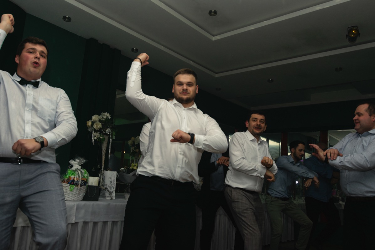 Grupa mężczyzn w białych koszulach tańczy na imprezie, jeden z nich ma założoną muchę, a na pierwszym planie widoczny jest stół z dekoracjami kwiatowymi i koszem prezentowym.