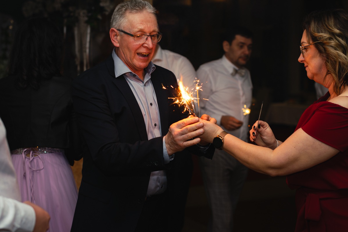 Uśmiechnięty mężczyzna w garniturze trzyma zapalone zimne ognie, przekazując je kobiecie w czerwonej sukience na tle gości weselnych.