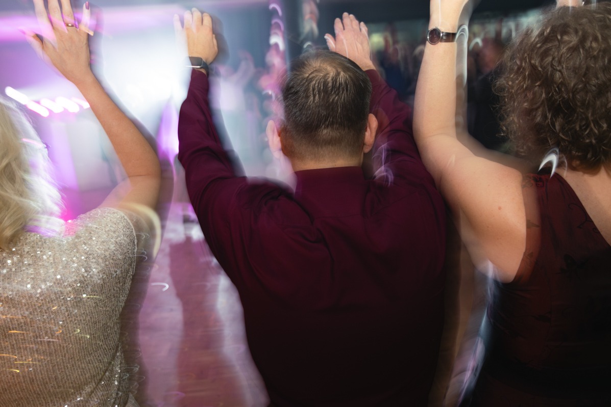 Rozmazane ujęcie trzech osób z podniesionymi rękami, prawdopodobnie podczas tańca na imprezie, widoczne błyszczące ubrania i oświetlenie dyskotekowe.