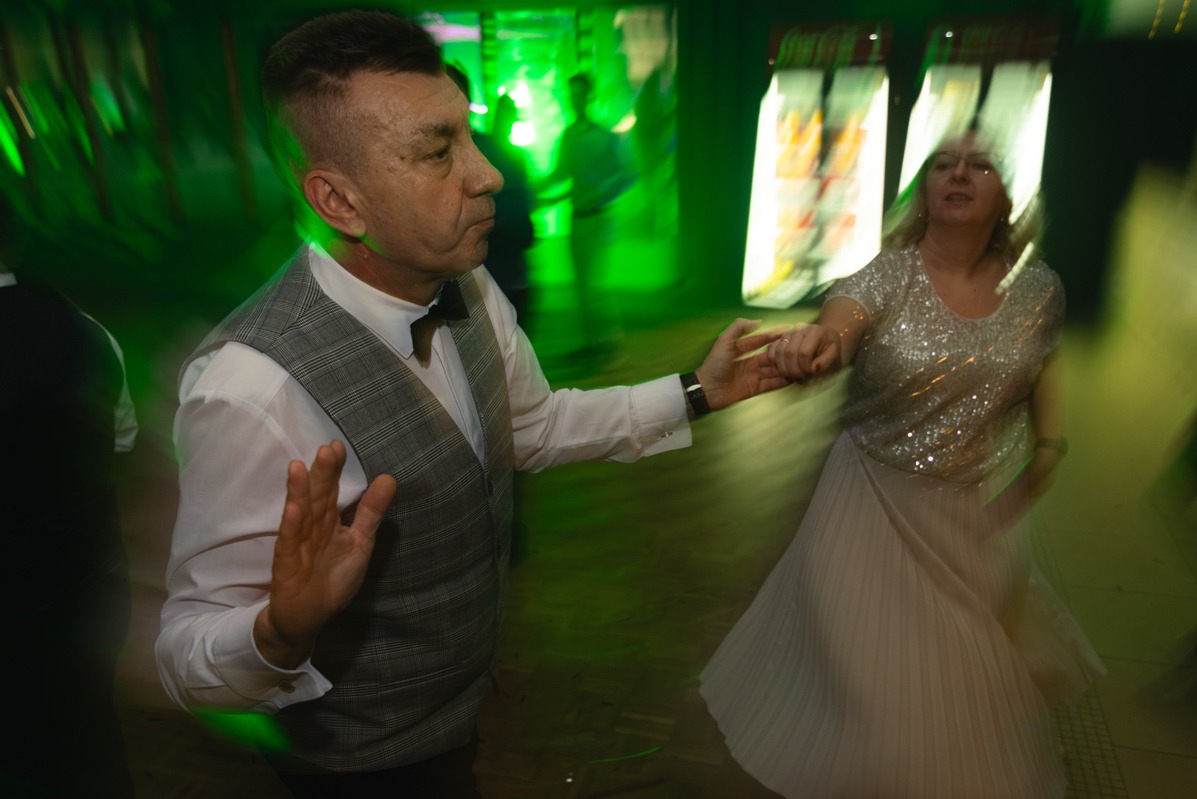 Para w ruchu tanecznym podczas wydarzenia, mężczyzna w kamizelce w kratę i muszce, kobieta w cekinowej bluzce, zielone oświetlenie sceniczne.