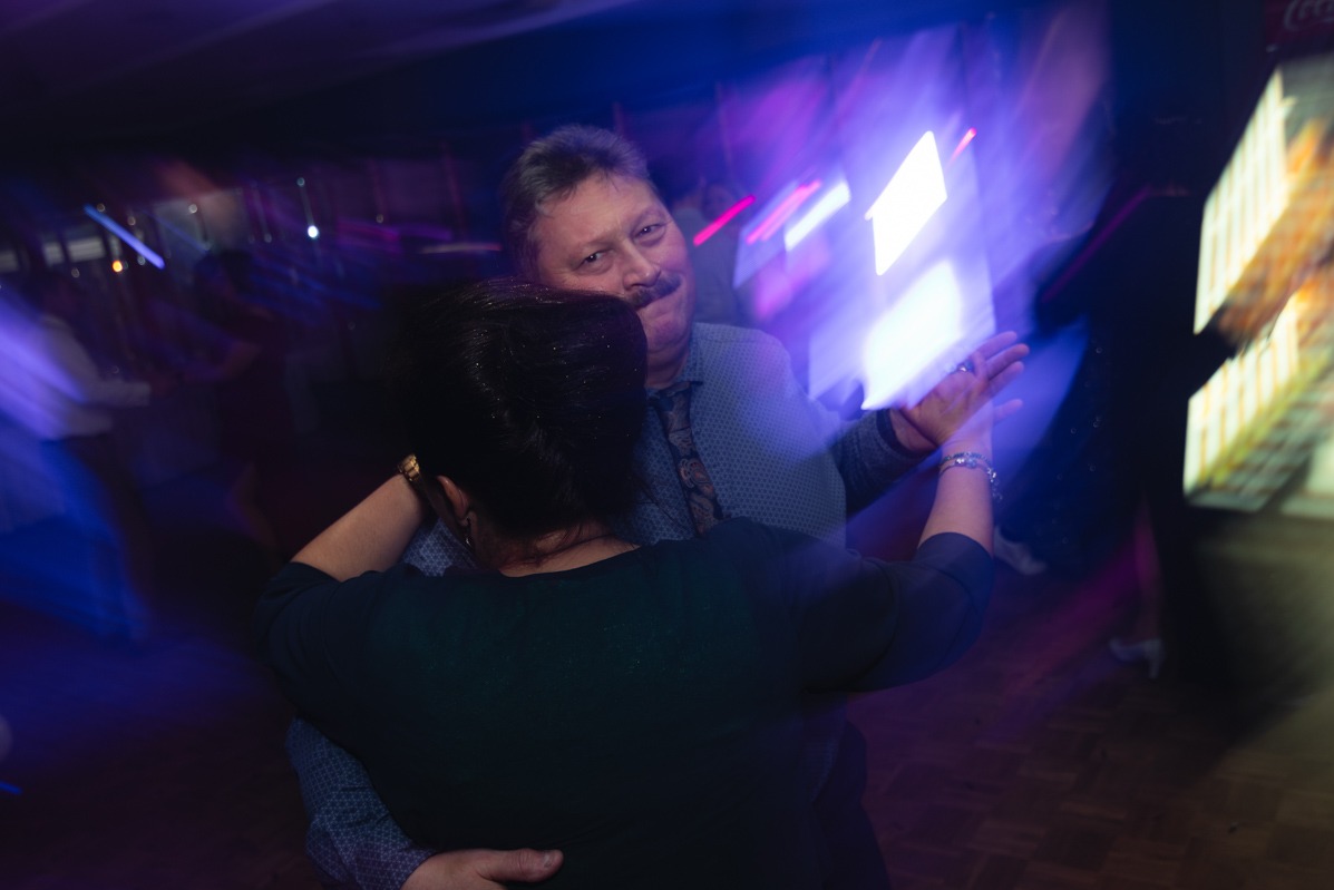 Para w średnim wieku tańczy na parkiecie podczas eventu, mężczyzna z wąsami w koszuli w drobny wzór i krawacie spogląda w obiektyw, oświetlenie dyskotekowe w tle.