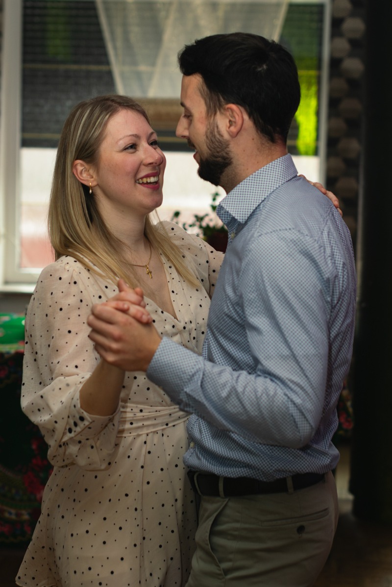 Uśmiechnięta para trzymająca się za ręce podczas tańca, kobieta w kremowej sukience w czarne kropki i złoty naszyjnik, mężczyzna w jasnoniebieskiej koszuli w drobną kratkę.