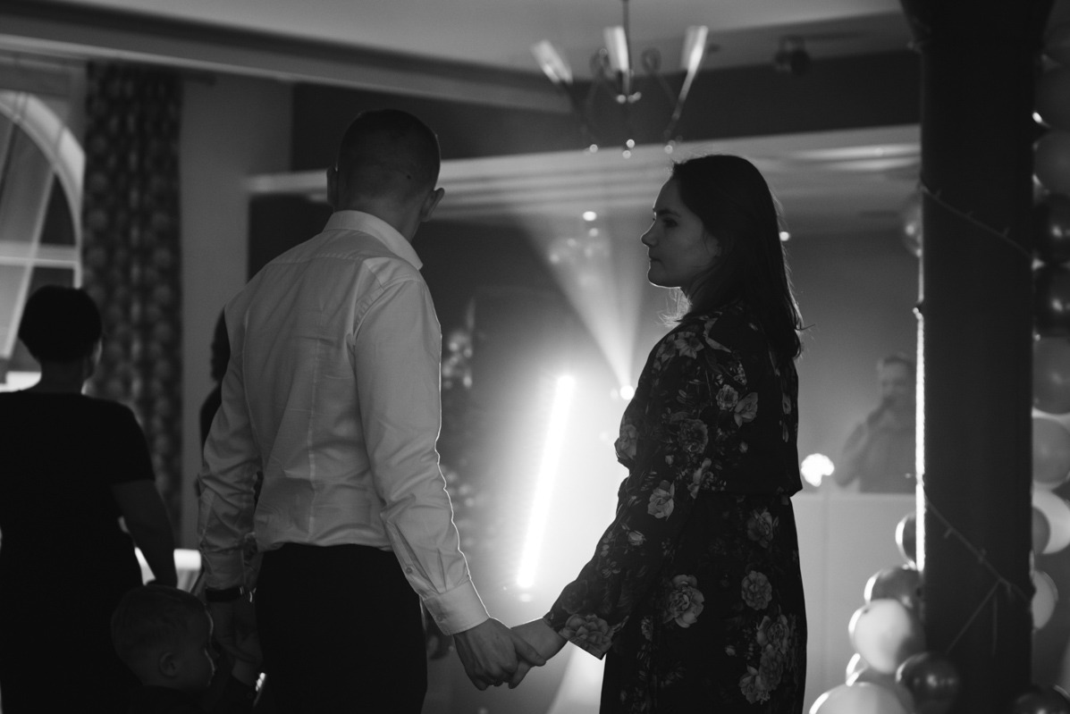 Czarno-białe zdjęcie pary trzymającej się za ręce na tle jasnych świateł i dekoracji balonowych, z widocznym DJ-em w tle.