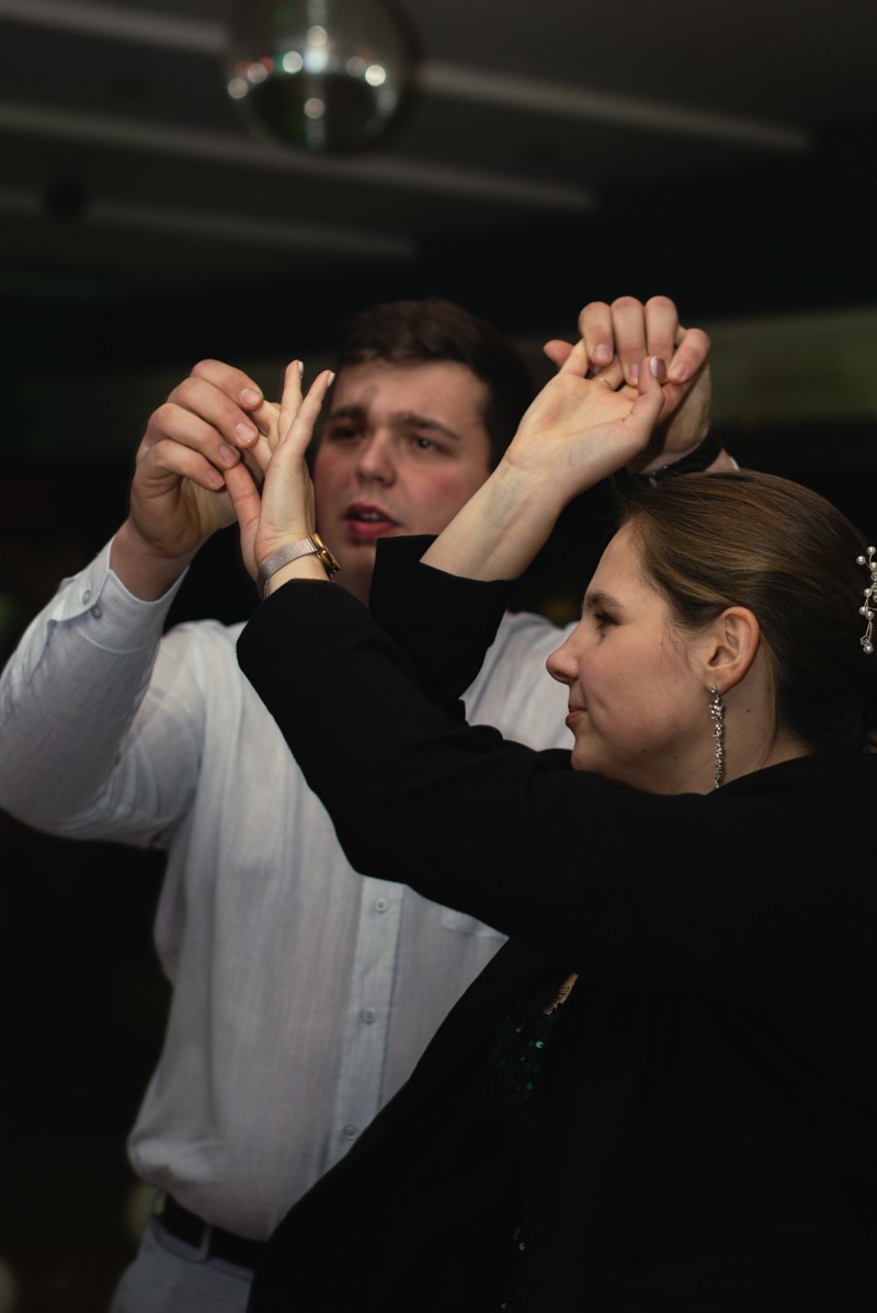 Para tańcząca na parkiecie pod dyskotekową kulą, mężczyzna w białej koszuli trzyma ręce kobiety w czarnym żakiecie.