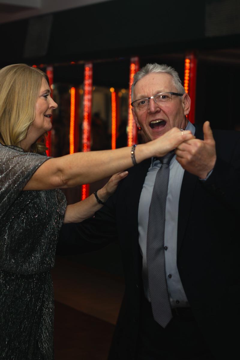 Para w średnim wieku tańczy na imprezie, mężczyzna w okularach z szeroko otwartymi ustami trzyma kobietę za rękę, w tle czerwone oświetlenie.