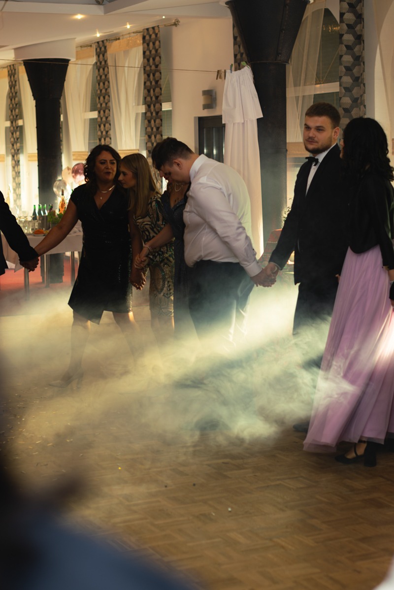 Grupa osób trzymających się za ręce na parkiecie w zadymionym pomieszczeniu podczas imprezy, widoczne dekoracje i oświetlenie