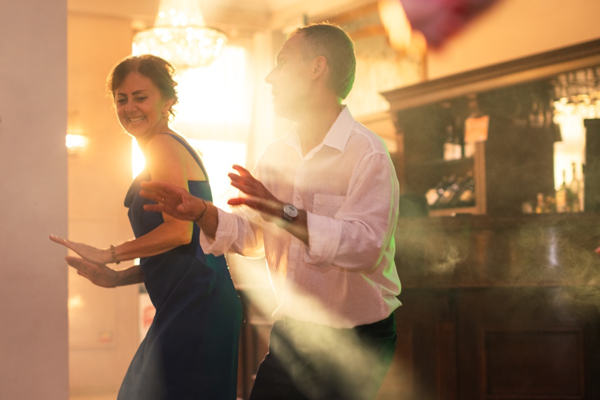 Uśmiechnięta kobieta w granatowej sukience i mężczyzna w białej koszuli tańczą podczas imprezy, oświetleni ciepłym światłem, z widocznym fragmentem baru w tle.