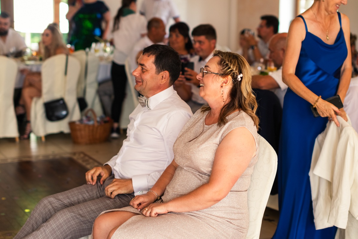 Uśmiechnięta para siedząca na krzesłach podczas uroczystości, mężczyzna w białej koszuli i muszce, kobieta w błyszczącej sukience z wpiętymi kwiatami we włosach, w tle goście i dekoracje.