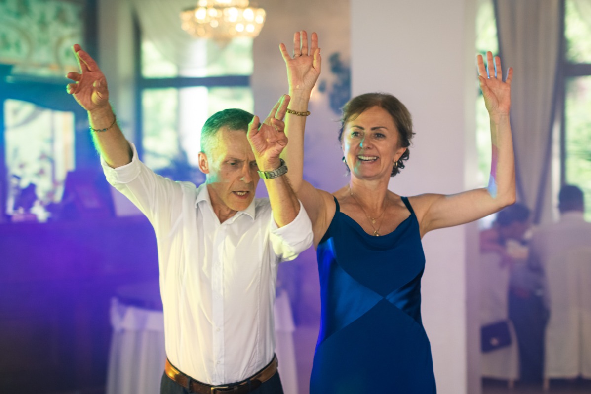 Para w średnim wieku tańcząca z podniesionymi rękami na sali balowej podczas imprezy, oświetlona niebieskim światłem.