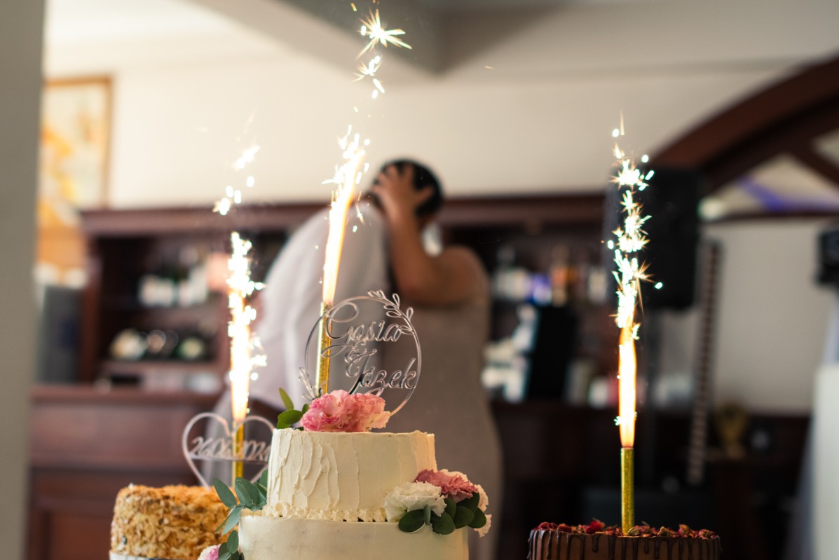 Trzy torty weselne z zimnymi ogniami, na środkowym tort z napisem 'Gosia & Jacek' i różowymi kwiatami, w tle całująca się para.