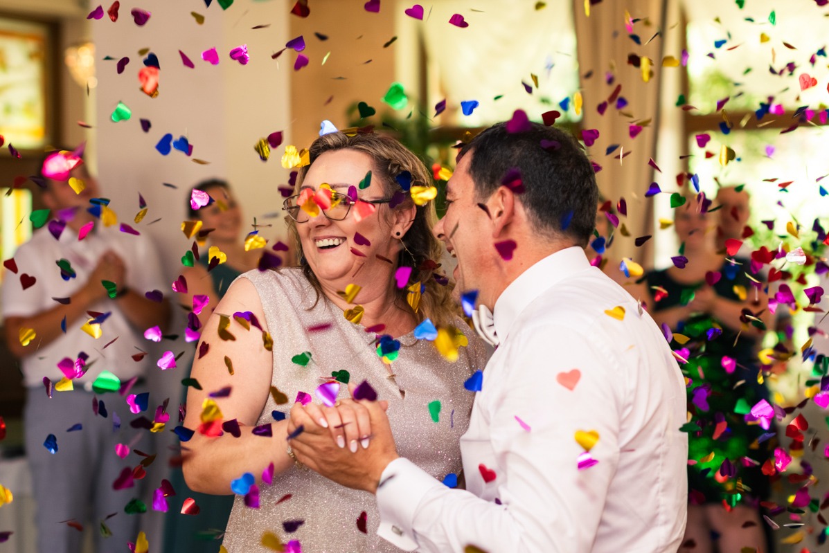 Para tańcząca w deszczu kolorowego konfetti w kształcie serc, kobieta w okularach z serduszkami, radosny wyraz twarzy, w tle uśmiechnięci goście.