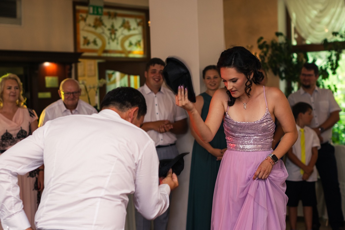 Panna młoda w liliowej sukni z cekinowym topem łapie rzucony kapelusz podczas oczepin na sali weselnej, w tle goście weselni.