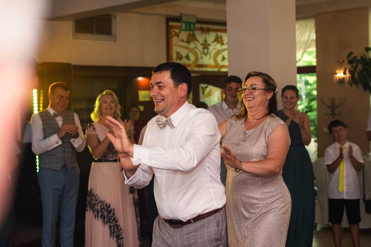 Para w średnim wieku, uśmiechnięta i klaszcząca w dłonie na sali balowej podczas uroczystości, otoczona przez innych gości również klaszczących.