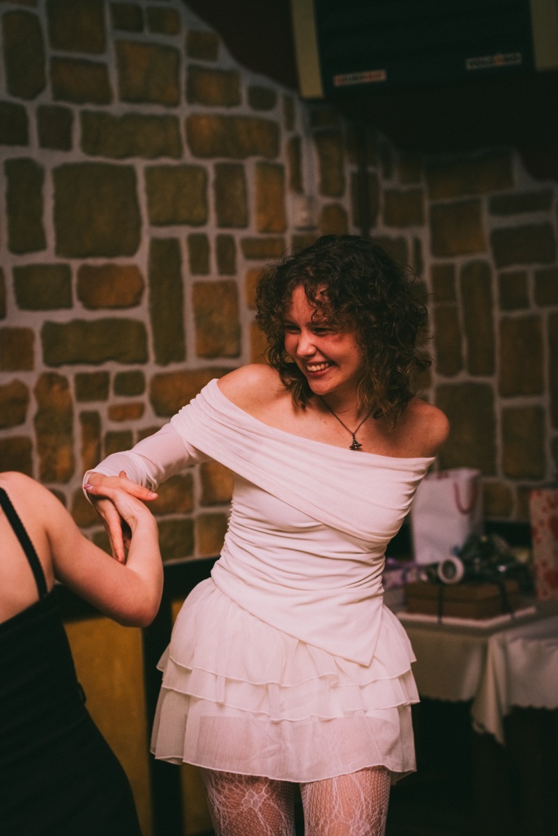 Uśmiechnięta kobieta w białej sukience z odkrytymi ramionami, trzymająca dłoń innej osoby, na tle ściany z kamienia podczas wydarzenia.