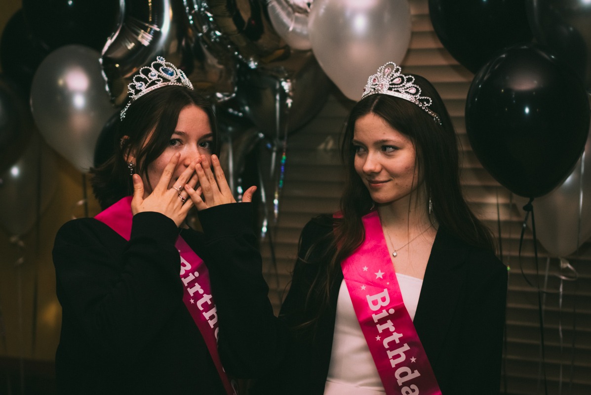 Dwie nastolatki w tiarach i szarfach urodzinowych na tle balonów w odcieniach bieli, srebra i czerni. Jedna dziewczyna zasłania usta dłońmi, druga patrzy w jej stronę z uśmiechem.