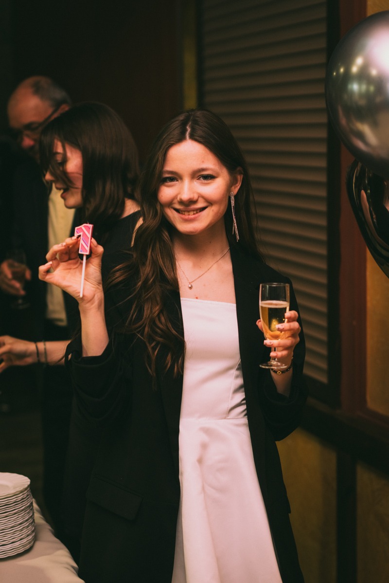 Uśmiechnięta młoda kobieta w białej sukience i czarnej marynarce trzyma świeczkę z numerem '1' i kieliszek szampana na tle imprezy urodzinowej.