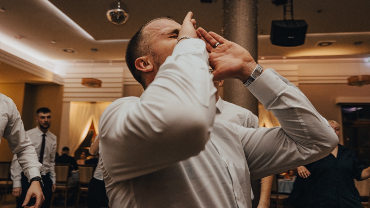 Mężczyzna w białej koszuli tańczy z rękami uniesionymi do góry podczas wydarzenia w sali z parkietem i lustrzaną kulą.