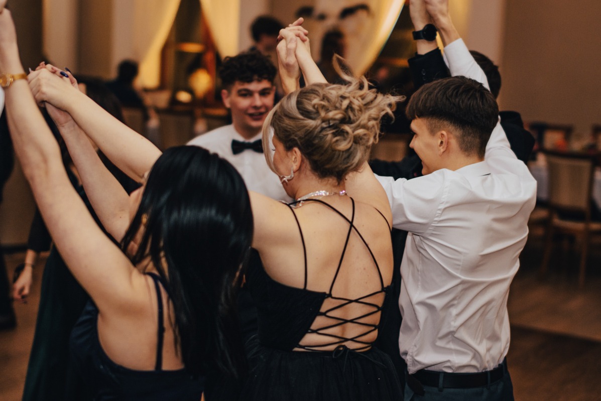 Grupa młodych ludzi, prawdopodobnie na balu maturalnym w Kędzierzynie-Koźlu, trzyma się za ręce w kręgu podczas tańca. Widoczna fryzura blond kobiety z tyłu oraz czarna sukienka z ozdobnymi...
