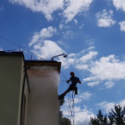 Grupa Doczyszczacze Monika Sosińska - Pracownik na linach, w kasku, czyści ścianę budynku strumieniem wody pod ciśnieniem; niebo z chmurami w tle.