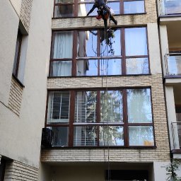 Grupa Doczyszczacze Monika Sosińska - Pracownik na linach zabezpieczających czyści elewację budynku wielorodzinnego, widoczne okna z brązowymi ramami i fragment elewacji z cegły, na dole drabina i rower.