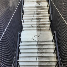 Grupa Doczyszczacze Monika Sosińska - Schody z granitu, w połowie wyczyszczone myjką ciśnieniową, widoczny kontrast między czystą a brudną powierzchnią, czarny kabel myjki zwinięty na schodach, metalowa balustrada po bokach.