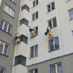 Trzech alpinistów przemysłowych maluje elewację bloku mieszkalnego, zabezpieczone okna folią i taśmą malarską, widoczne liny i wiadra z farbą.