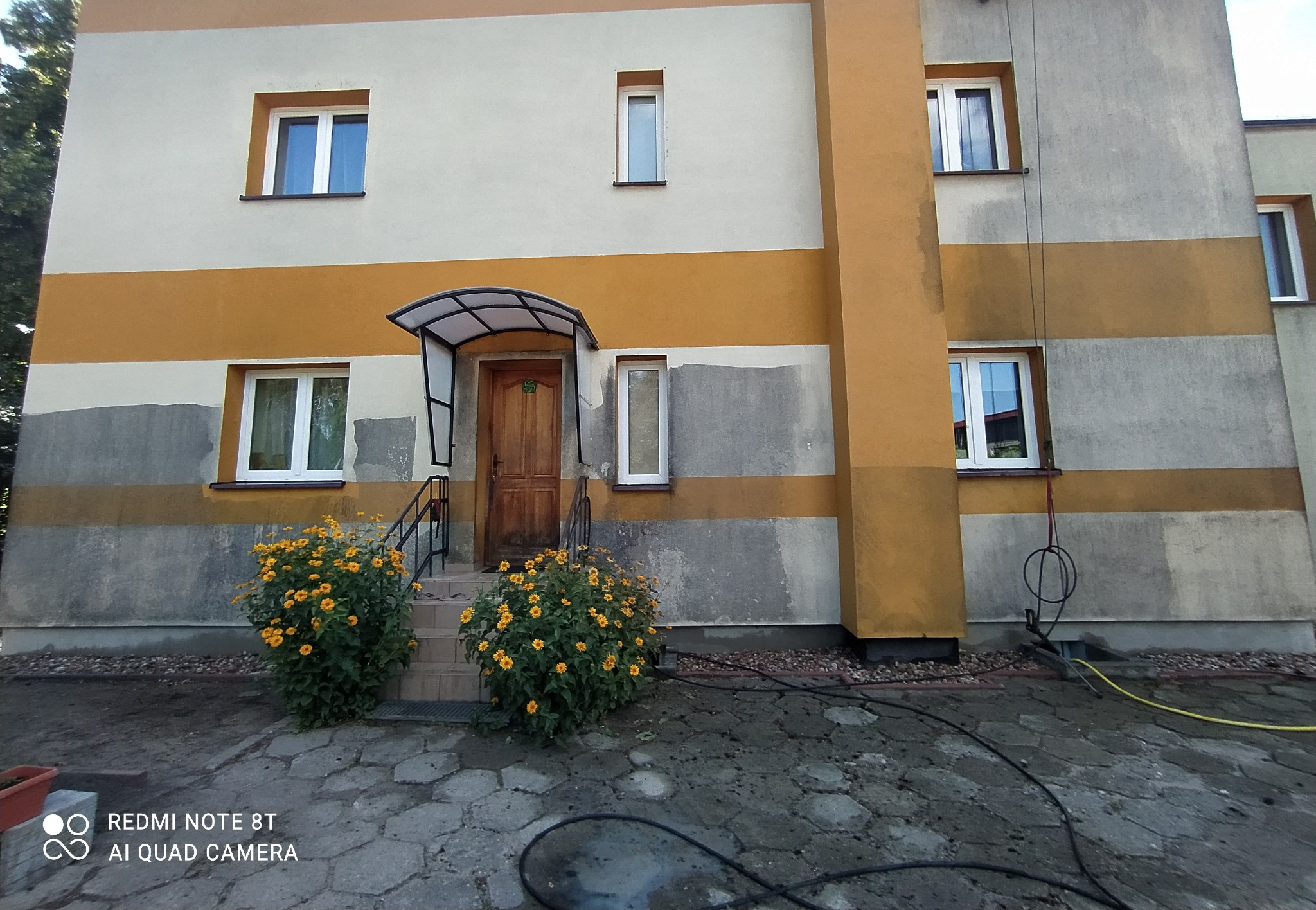 Dwukondygnacyjny budynek z częściowo odnowioną elewacją, widoczne ślady niedoczyszczonych fragmentów i węże do mycia na chodniku, przed wejściem kwitnące krzewy.