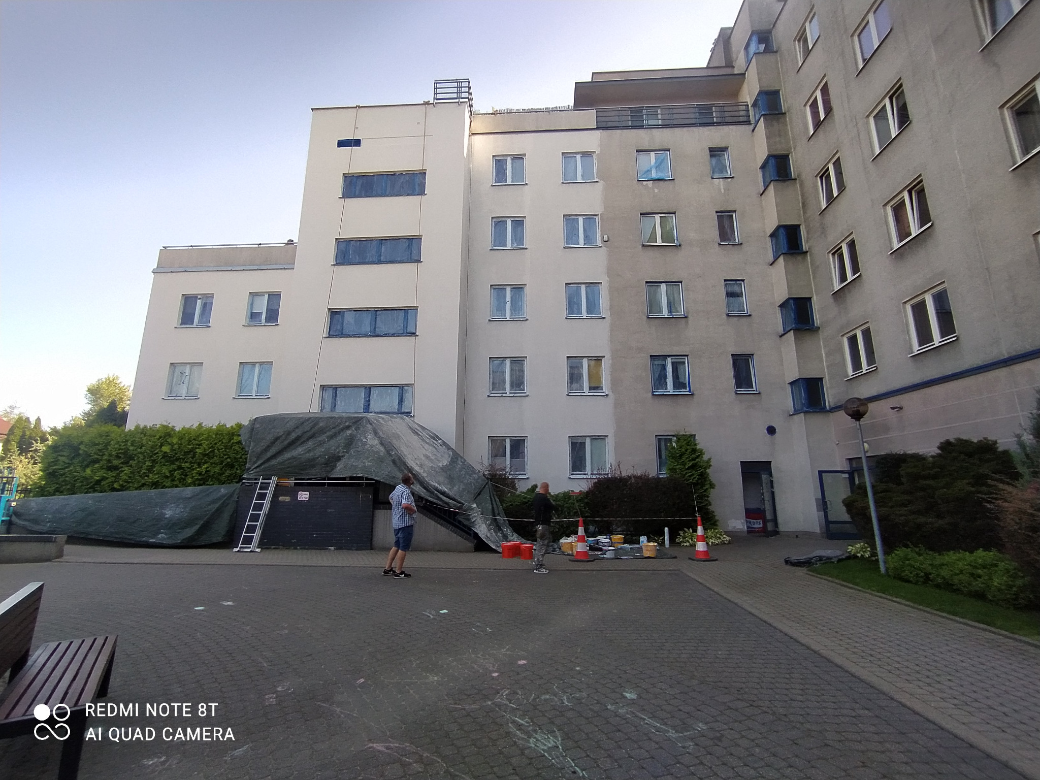 Wielopiętrowy budynek z częściowo odnowioną elewacją, widoczne przygotowania do prac: rozłożona plandeka, drabina, wiadra z farbą i stożki ostrzegawcze, dwóch mężczyzn w trakcie pracy.