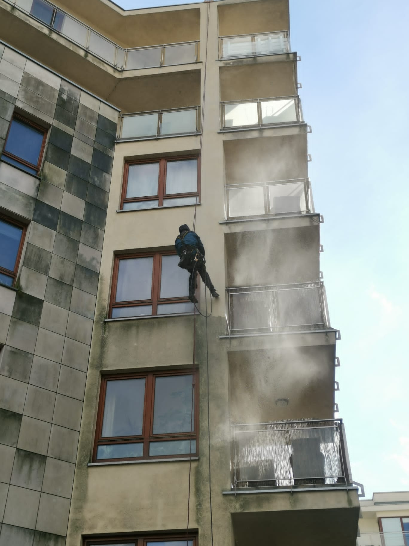 Pracownik na linie, w uprzęży, czyści elewację budynku wielorodzinnego pod ciśnieniem, widoczne ślady po czyszczeniu na balkonach i ścianie.