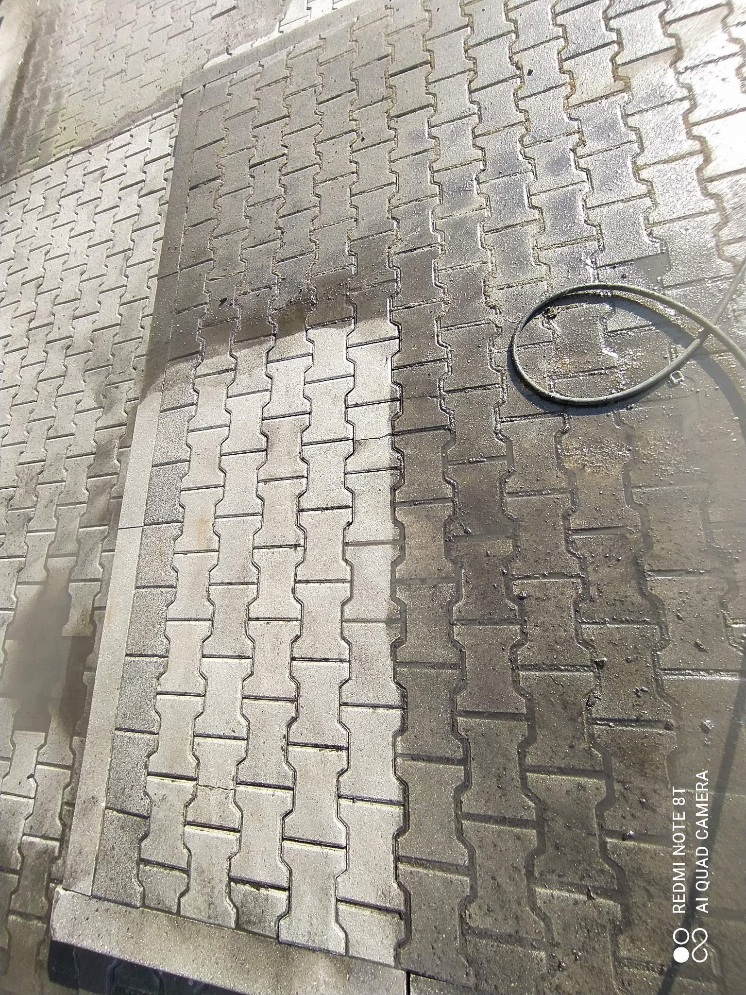 Częściowo wyczyszczona kostka brukowa z widoczną różnicą w kolorze między umytą a brudną powierzchnią, obok leży zwinięty wąż.