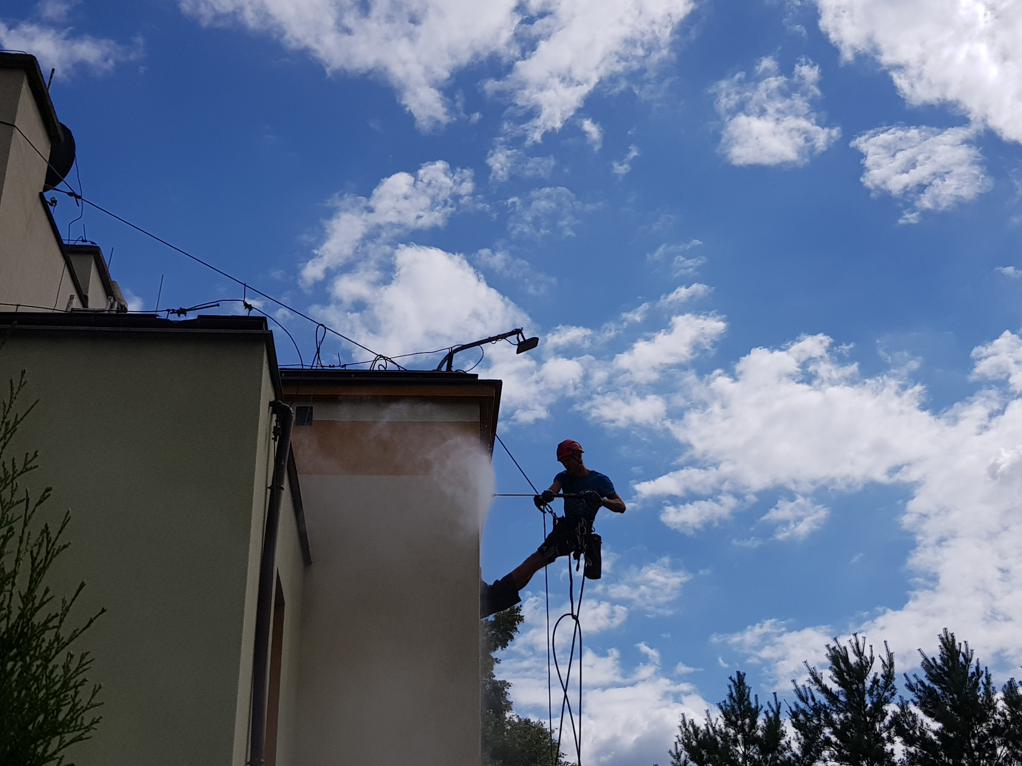 Pracownik na linach, w kasku, czyści ścianę budynku strumieniem wody pod ciśnieniem; niebo z chmurami w tle.