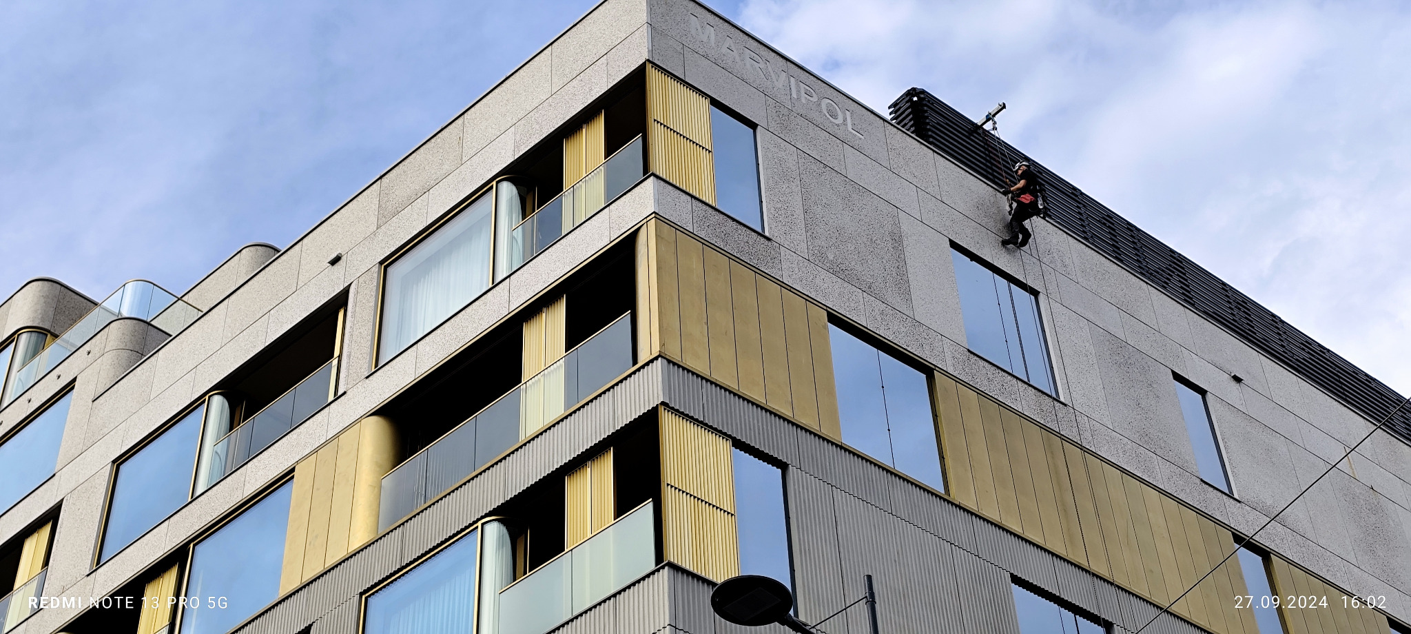 Pracownik na linach zabezpieczających wykonuje prace na elewacji nowoczesnego budynku z elementami w kolorze złota i dużymi oknami, widoczny napis 'MARVIPOL' na elewacji.