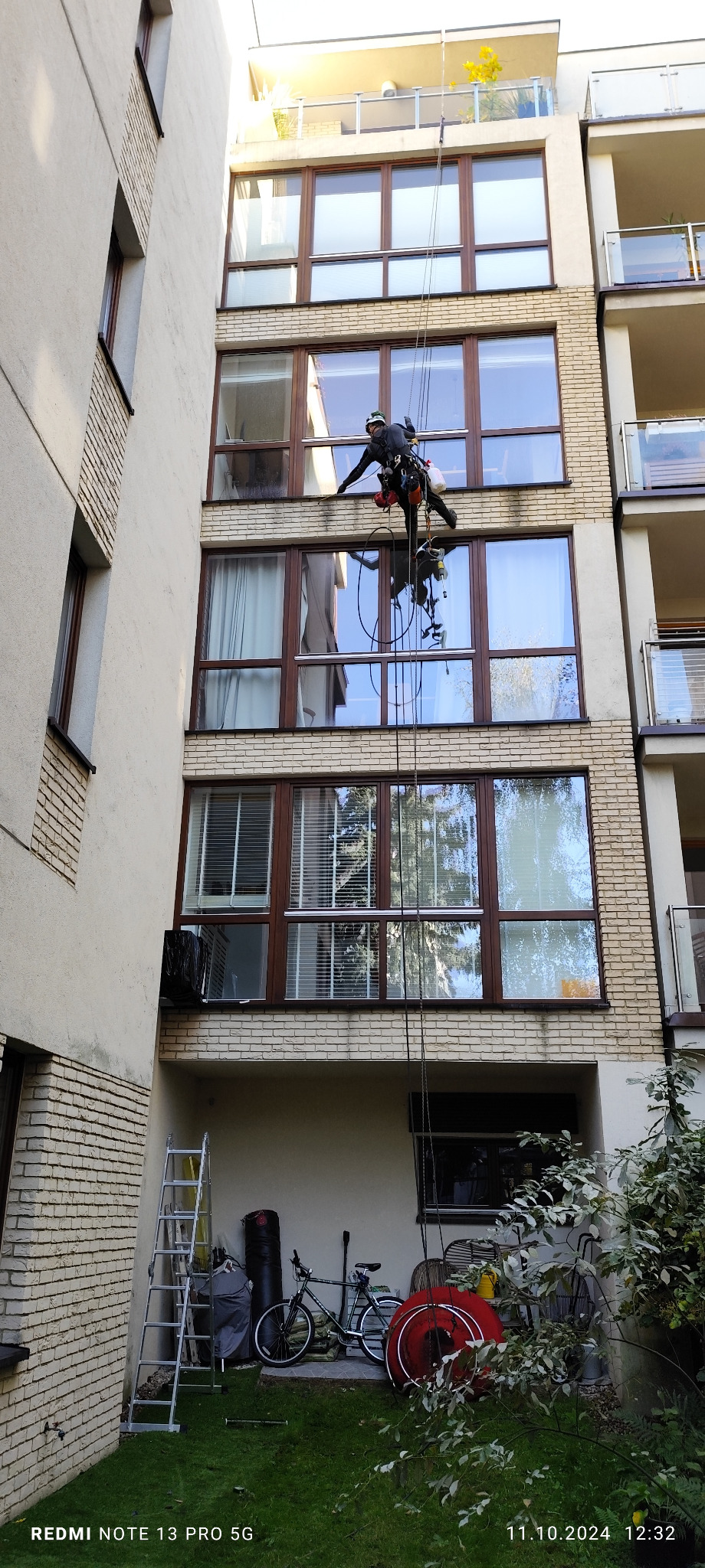 Pracownik na linach zabezpieczających czyści elewację budynku wielorodzinnego, widoczne okna z brązowymi ramami i fragment elewacji z cegły, na dole drabina i rower.
