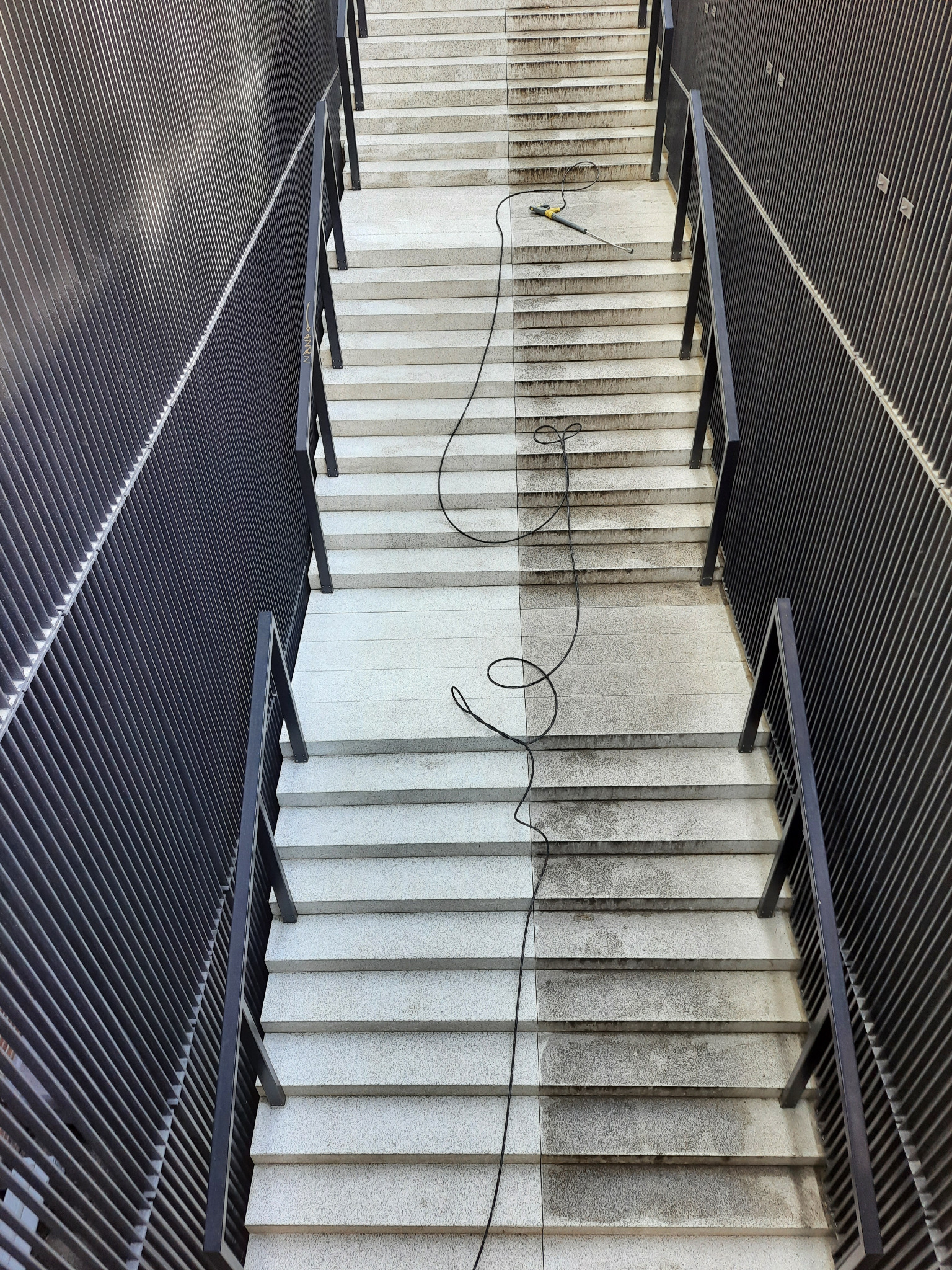 Schody z granitu, w połowie wyczyszczone myjką ciśnieniową, widoczny kontrast między czystą a brudną powierzchnią, czarny kabel myjki zwinięty na schodach, metalowa balustrada po bokach.