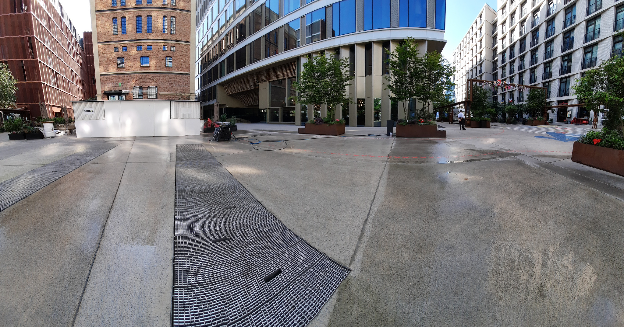 Szeroki kadr na plac miejski z betonową nawierzchnią, czyszczoną za pomocą myjki ciśnieniowej, z widocznymi budynkami o nowoczesnej architekturze i elementami zieleni w donicach.