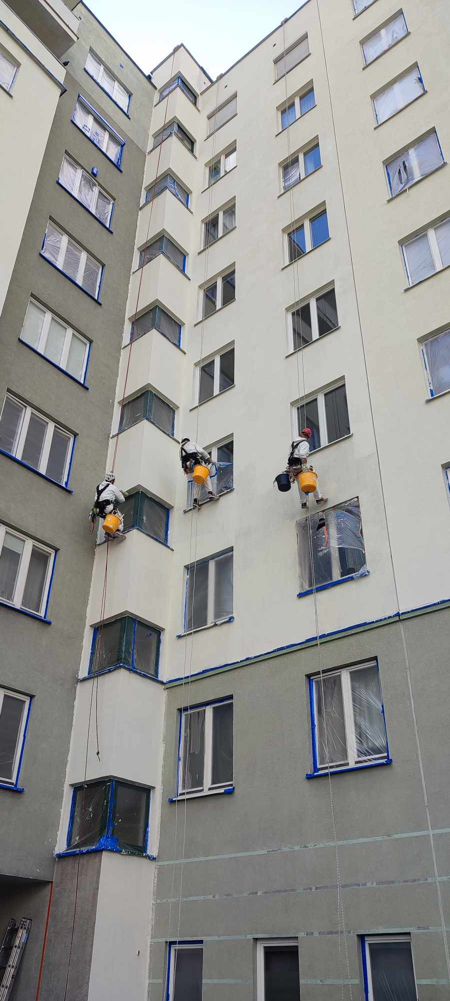 Trzech alpinistów przemysłowych maluje elewację bloku mieszkalnego, zabezpieczone okna folią i taśmą malarską, widoczne liny i wiadra z farbą.