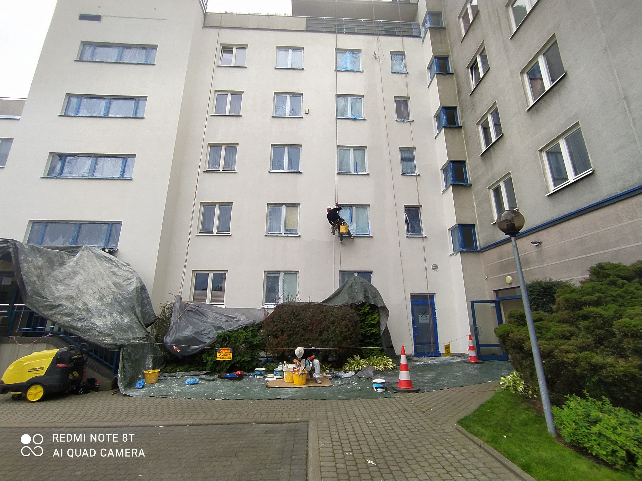 Pracownik na linie zabezpieczonej, maluje elewację budynku wielorodzinnego, okna zabezpieczone folią, na dole rozłożona plandeka i przygotowane farby.