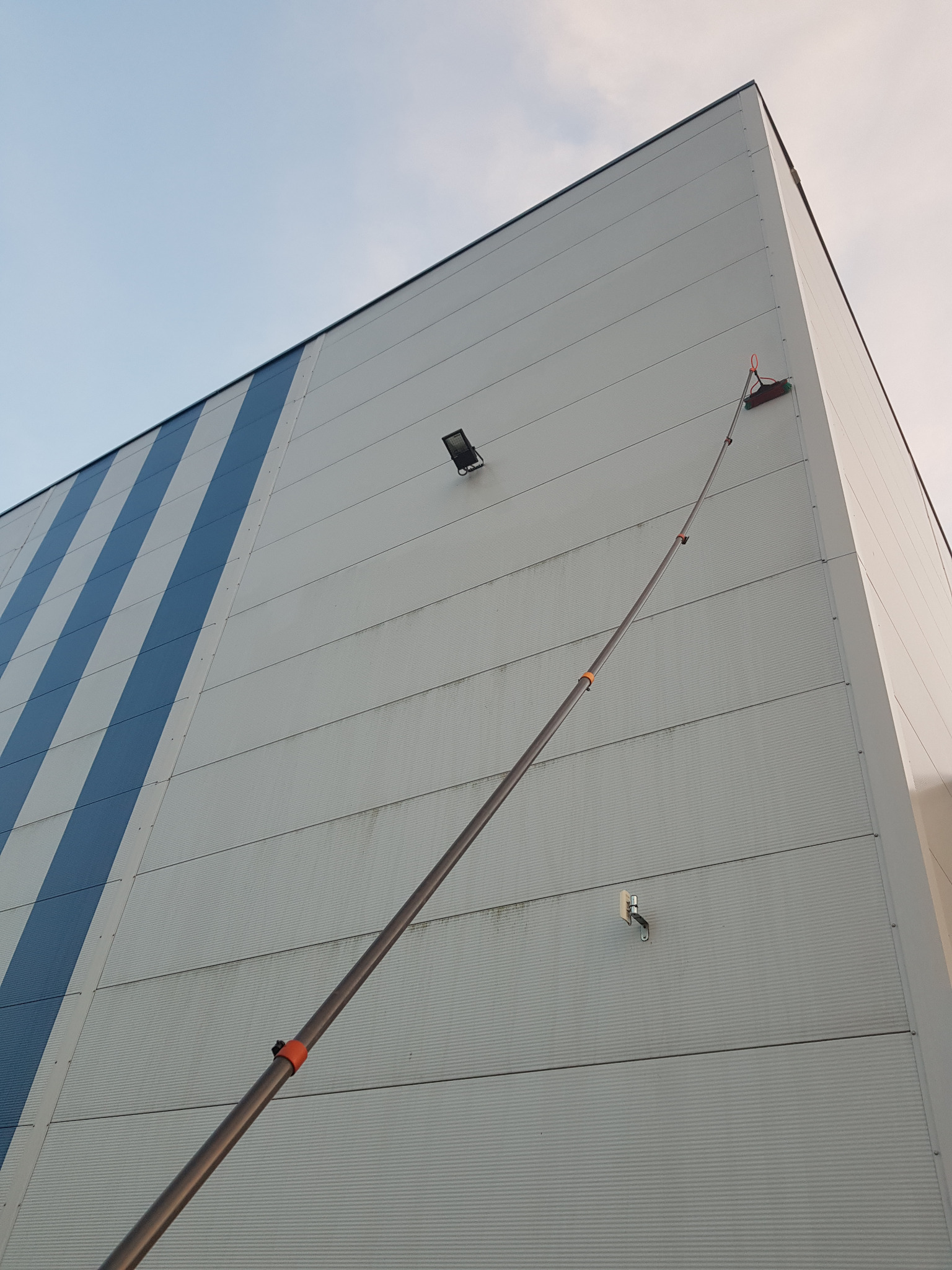 Wysoka ściana budynku przemysłowego z niebieskimi pasami, czyszczona za pomocą szczotki na długim teleskopowym kiju, widoczna lampa oświetleniowa i element mocujący na elewacji.