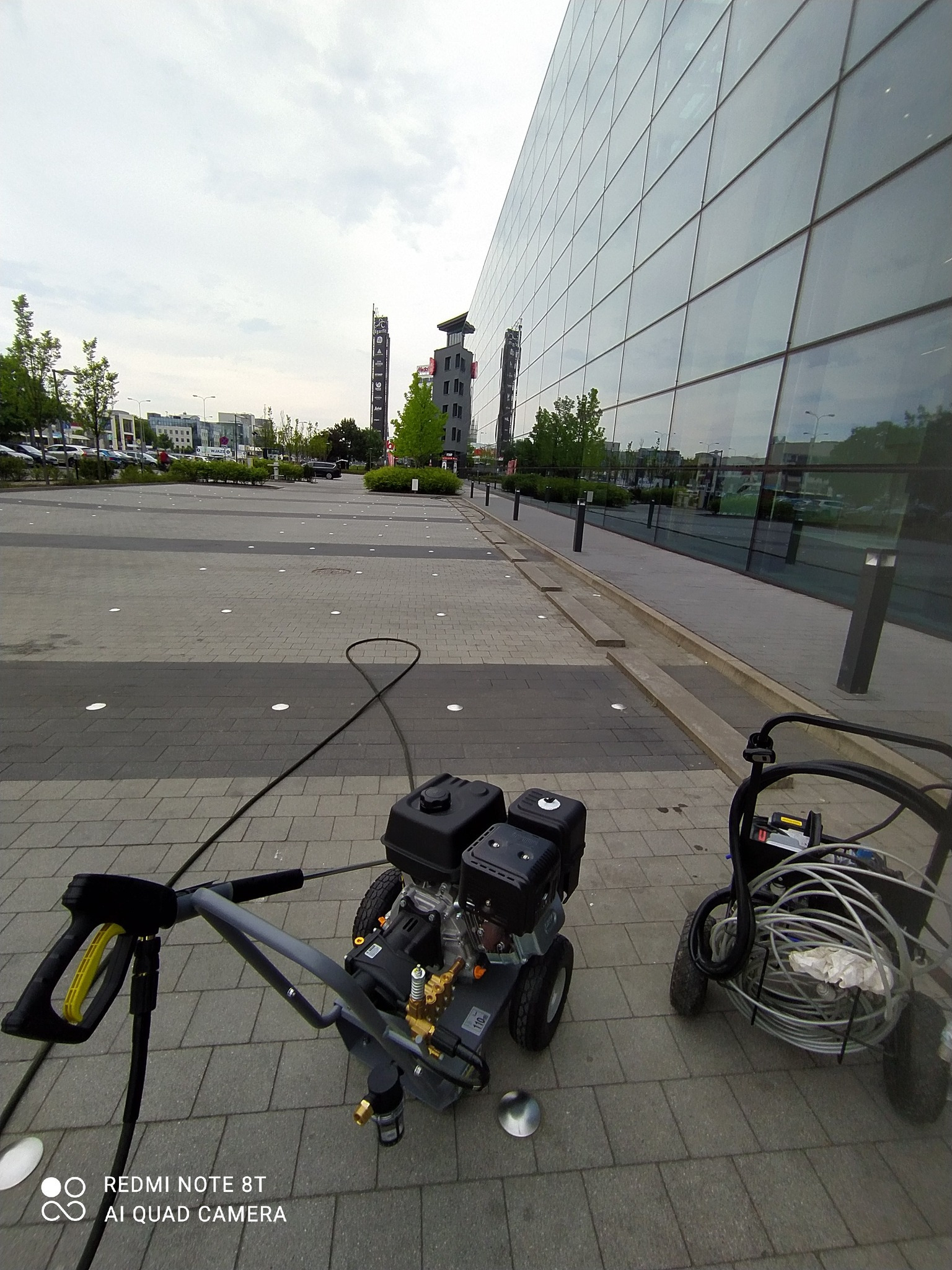 Dwa urządzenia wysokociśnieniowe na kostce brukowej przed nowoczesnym biurowcem z szklaną fasadą. W tle widoczne drzewa i budynki miejskie.